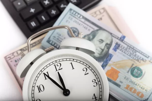 A close-up image showing a classic analog alarm clock foreground with its hands indicating a time close to 2 o'clock, placed next to a blurred stack of US hundred-dollar bills and a calculator in the background, symbolizing the concept of Early Wage Access.